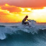 Surfer mit Surfer-Sonnenbrilleauf einer Welle mit Sonnenuntergang im Hintergrund