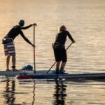 Zwei Personen betreiben Stand-Up-Paddling auf dem Meer bei Sonnenuntergang und tragen dabei SUP Socken.