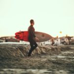 Ein Mann im Neoprenanzug trägt ein SUP-Board. Dies kann mit einer SUP Elektropumpe aufgepumpt werden.