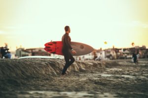 Ein Mann im Neoprenanzug trägt ein SUP-Board. Dies kann mit einer SUP Elektropumpe aufgepumpt werden.