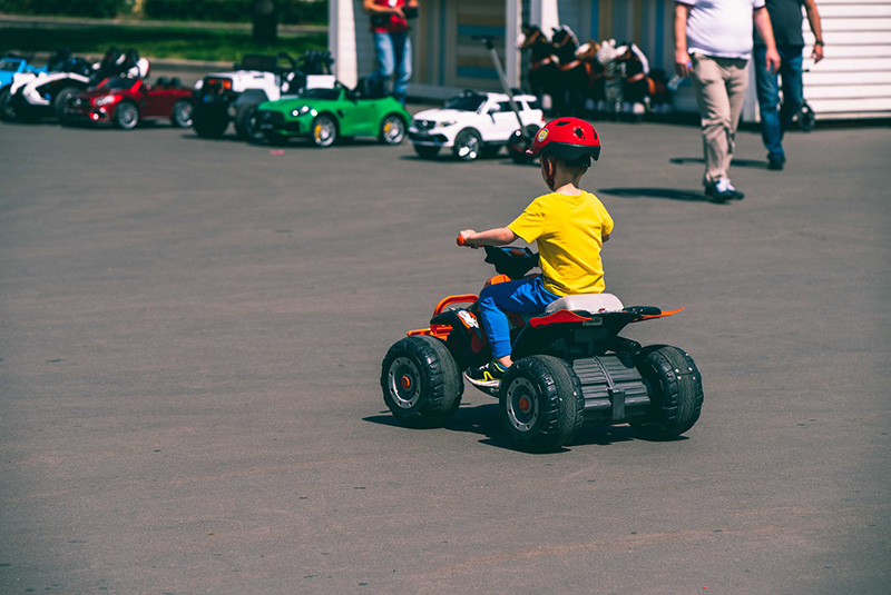 Kleiner Junge, Fahrer eines elektrischen Quads für Kinder mit gelben T-Shirt fährt auf Asphalt.