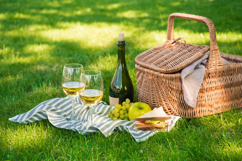 Picknickkorb auf einer Wiese, sowie gefüllte Weingläser, eine Weinflasche und Speisen auf einer Decke ausgebreitet