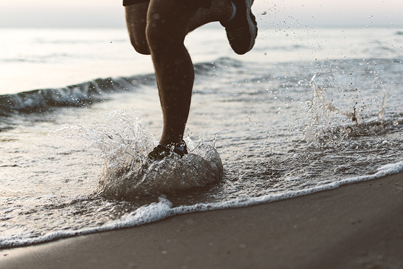 Die Beine einer laufenden Person, die mit ihren Schuhen durch die Brandung des Meeres am Strand läuft.