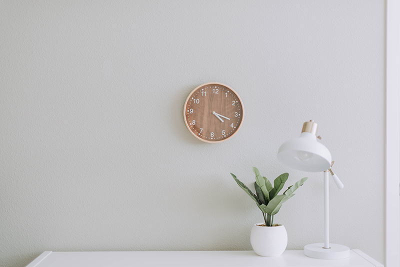 Ein minimalistisches Bild mit weißer Wand, an der eine braune Uhr in Holzoptik hängt. Auf der Kommode, von der wir nur einen sehr kleinen Ausschnitt der Ablagefläche sehen können, stehen zur Rechten eine weiße Vase mit einer Grünpflanze, sowie daneben eine weiße Tischlampe.