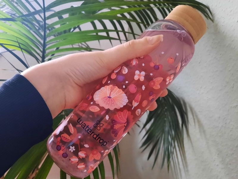 Hand hält eine mit floralen Mustern bedruckte Waterdrop-Flasche vor einer Pflanze.