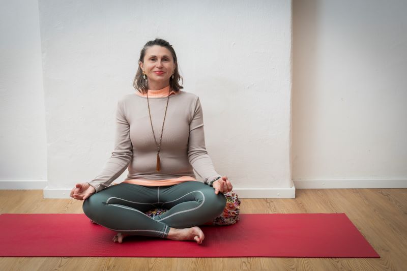 Eine Frau sitzt in einer Meditationshaltung auf einer roten Yogamatte in einem Yoga-Raum,